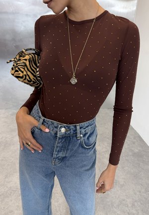 Personne portant un top marron à manches longues avec des pois dorés, un jean bleu taille haute, un long collier pendentif et tenant un petit sac à main à rayures de tigre.