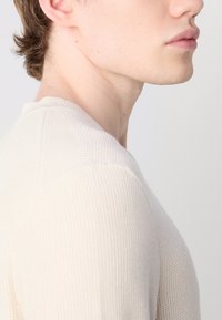 Maglione a coste di un beige chiaro, con scollatura tonda e una sottile texture. Presenta un design fitted e cuciture visibili sulle spalle.
