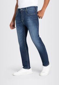 Jeans bleu foncé ajustés, dotés d'un design classique à cinq poches, de coutures contrastantes et d'une fermeture à bouton. Portés avec des baskets blanches.