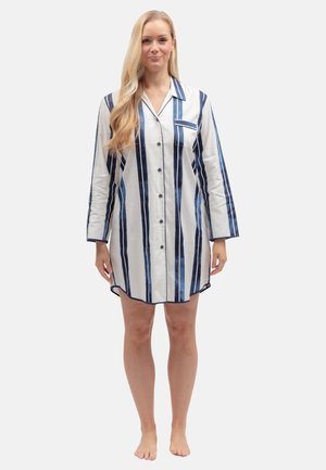 Femme debout pieds nus portant une robe chemise blanche à manches longues avec des rayures verticales bleues et une poche poitrine.