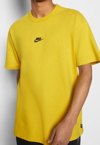 Žluté tričko Nike vyrobené z bavlny, s černým vyšívaným logem na hrudi, krátkými rukávy a volným střihem.