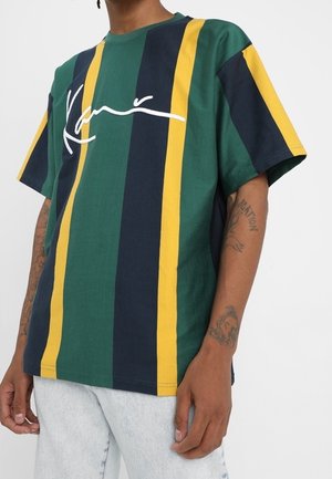 Personne portant un t-shirt rayé ample avec des bandes verticales vertes, bleu marine et jaunes, et un texte blanc en écriture cursive sur la poitrine, associé à un jean en denim clair.