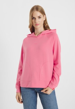 Vrouw die een felroze hoodie met zakken aan de voorkant en een blauwe spijkerbroek draagt, staand tegen een effen lichte achtergrond.