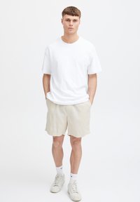 Tricou alb cu mâneci scurte, asortat cu pantaloni scurți bej cu textură. Pantofi sport albi și șosete până la gleznă completează ținuta casual. Fundal neutru.