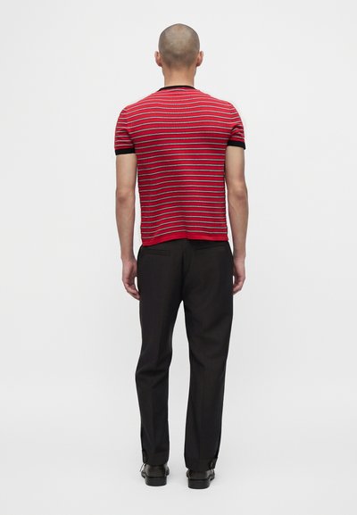 Homme debout, de dos, portant une chemise à manches courtes rayée rouge et noire, pantalon noir et chaussures noires sur fond blanc.