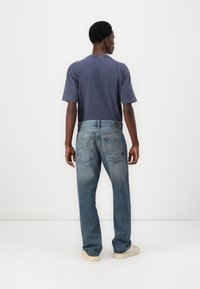 Mėlynos denim kelnės su laisvu kirpimu, penkiomis kišenėmis ir švelniu blukimu. Sukomplektuota su tamsiai mėlynu marškinėliu, matoma iš nugaros.