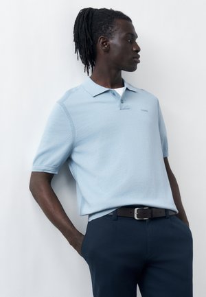 Mann mit Dreadlocks, der ein hellblaues kurzärmeliges Poloshirt und dunkelblaue Hose trägt, steht mit den Händen in den Taschen und schaut zur Seite.