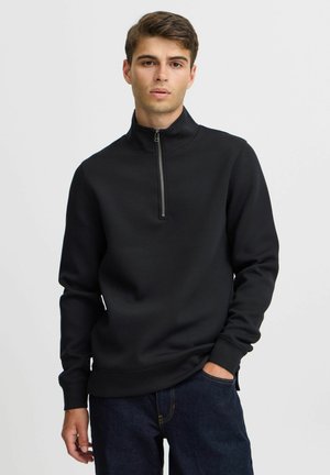 Schwarzer Zip-Hoodie aus glattem Stoff mit hohem Kragen und gefütterten Ärmeln. Kombiniert mit dunkelblauen Jeans.
