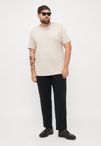 Beige katoenen t-shirt met korte mouwen en een klein logo. Gecombineerd met zwarte broek en zwarte schoenen. Model draagt een zonnebril.