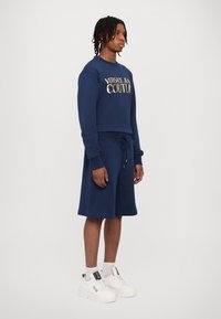 Mornarsko modra bombažna pulover s kapuco z zlatim logotipom "VERSACE JEAN COUTURE", v kombinaciji z ujemajočimi se kratkimi hlačami in belimi športnimi copati, ki stoji na sivi ozadju.
