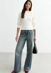 Maglione crema con dettaglio a fiocco al collo, abbinato a jeans a gamba larga blu chiaro e scarpe bordeaux, mentre tiene una piccola borsa nera.