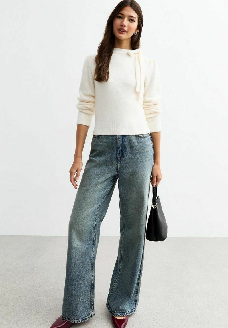 Maglione crema con dettaglio a fiocco al collo, abbinato a jeans a gamba larga blu chiaro e scarpe bordeaux, mentre tiene una piccola borsa nera.
