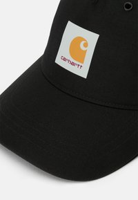 Crna kapa s ravnim obodom i teksturom od tkanine. Ima kvadratnu bijelu flaster s žutim logotipom i "carhartt" ispisano crvenim slovima.