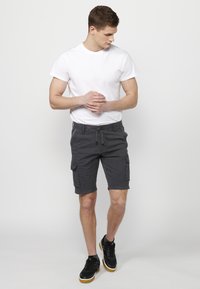 Ung man i vit t-shirt och mörka cargo-shorts som står barfota på ett ben, tittar ner med händerna i kors.