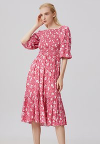 Robe florale rose avec un corsage smocké, des manches bouffantes et une jupe à volants. Le tissu a une texture douce avec des motifs de fleurs blanches et jaunes.
