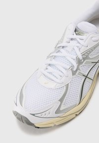 Weißer Sportschuh mit Obermaterial aus Mesh, silbernen Akzenten, runder Zehenpartie und strukturiertem Sohlenprofil. Verfügt über Schnürsenkel und einen gepolsterten Kragen für zusätzlichen Komfort.