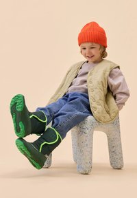 Grüne und schwarze isolierte Stiefel mit hellgrünen Akzenten und strukturierten Sohlen. Getragen von einem Kind in einer beigen Steppweste und einer orangefarbenen Mütze.