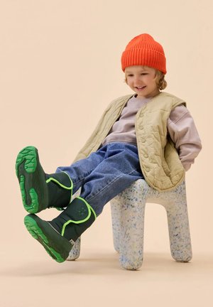 Grüne und schwarze isolierte Stiefel mit hellgrünen Akzenten und strukturierten Sohlen. Getragen von einem Kind in einer beigen Steppweste und einer orangefarbenen Mütze.