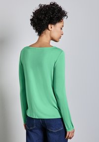 Długi rękaw, miętowa bluzka z szerokim dekoltem w łódkę i dopasowanymi mankietami, wykonana z gładkiego, miękkiego materiału. Zestawiona z ciemnoniebieskimi dżinsami.