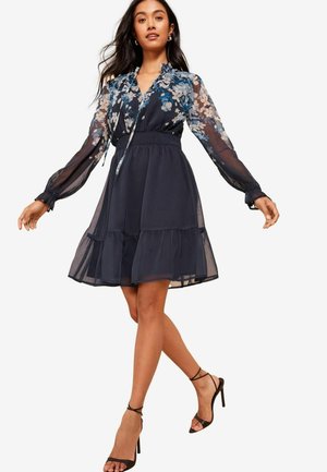 Navy blue dress with floral pattern, sheer long sleeves, smocked waist, layered skirt, and paired with black strappy high heels.