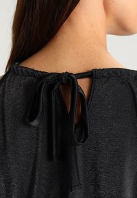 Černý top s texturovaným metalickým povrchem. Má klíčovou otvor na zadní straně s vázáním a jemný, řasený výstřih.