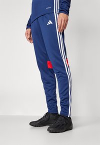 Navy athletic pants with white side stripes, red pocket accents, and ribbed cuffs, made from moisture-wicking fabric. Black sports shoes visible.