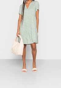 Vestido verde de manga corta con escote en V, con un patrón de hojas. La modelo sostiene un bolso tote beige y lleva sandalias trenzadas de color claro.