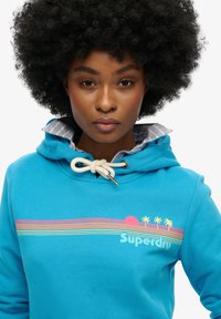 Turquoise hoodie met trekkoorden en een grafisch ontwerp met strepen, zon en palmbomen. Zachte textuur, comfortabele pasvorm, gelaagde kraag.
