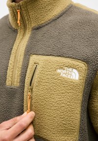 Primer plano de una mano cerrando con cremallera el bolsillo de forro polar color canela en una chaqueta North Face de forro polar gris y canela.