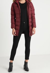 Chaqueta acolchada burdeos con capucha, cierre frontal con cremallera, textura acolchada, combinada con pantalones ajustados negros y botines.