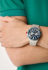 Chronografické hodinky s námořnickou modrou ciferníkem, stříbrným nerezovým páskem, červeným akcentačním tlačítkem a dvěma bílými chronografickými subciferníky.