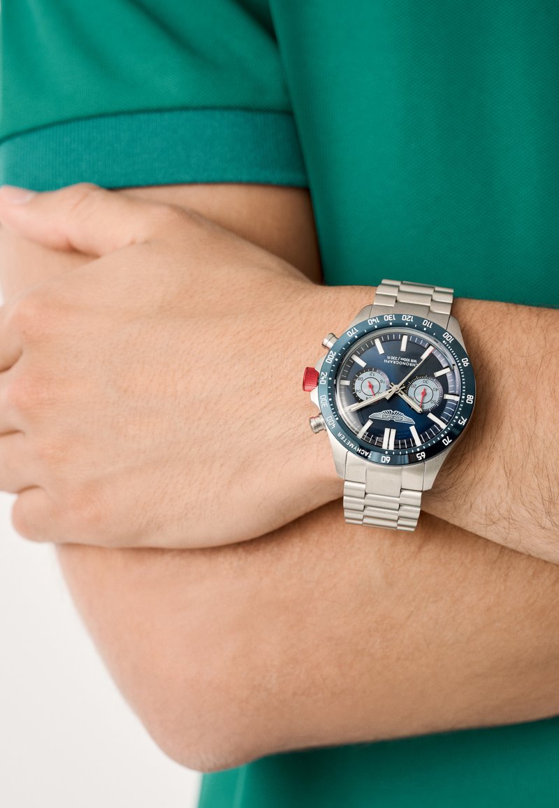 Chronografické hodinky s námořnickou modrou ciferníkem, stříbrným nerezovým páskem, červeným akcentačním tlačítkem a dvěma bílými chronografickými subciferníky.
