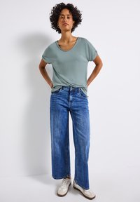 T-shirt oversize vert clair avec un large col, associé à un jean en denim bleu ample et des baskets blanches. Tissu lisse, style décontracté.