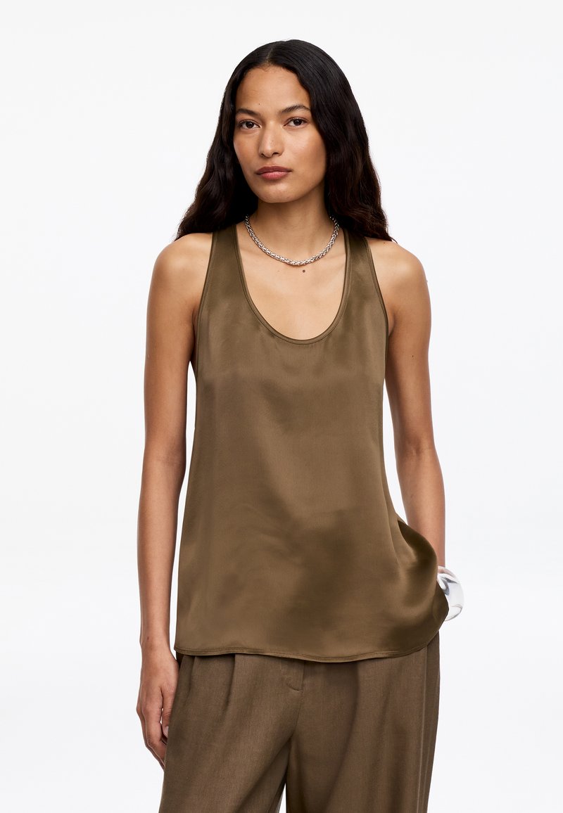 Femme aux longs cheveux foncés portant un haut en satin olive sans manches et un pantalon large assorti, accessoirisée d'un collier et d'un bracelet en chaîne.