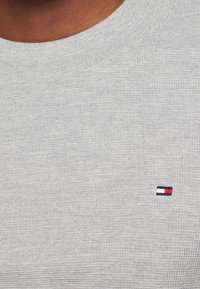 Harmaa neulepaita, jossa on pyöreä pääntie ja teksturoitu kangas; siinä on pieni brodeerattu logo punaisena, valkoisena ja sinisenä rinnan kohdalla.