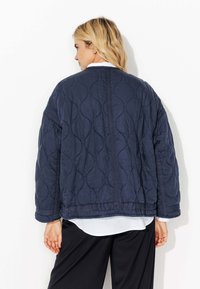 Veste matelassée bleu marine avec un motif de vagues texturé, offrant une coupe décontractée et des manches larges, superposée sur une chemise blanche.