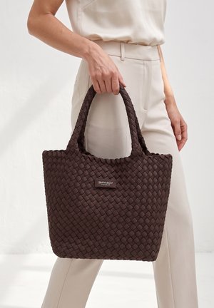 Personne portant une tenue beige tenant un sac cabas tissé brun foncé avec des poignées courtes et une étiquette "DIANA&CO FIRENZE".