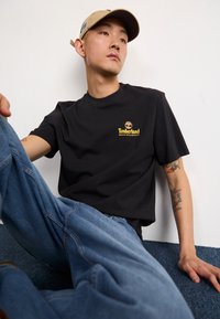 Camiseta de algodón negra con el logo amarillo de Timberland, combinada con jeans de mezclilla azul claro. Modelo sentado contra un fondo azul texturizado.