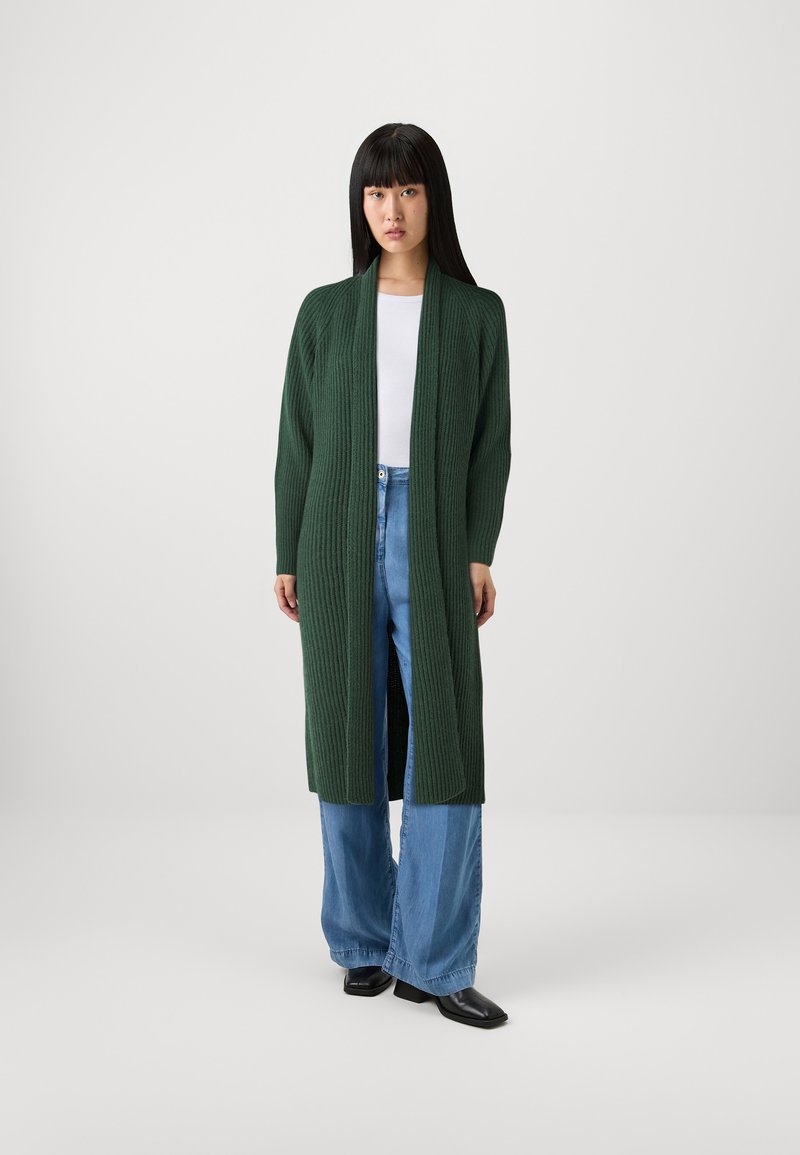 Cardigan lungo verde scuro con una texture a coste, apertura frontale e spacco laterale, indossato sopra una maglietta bianca e jeans a gamba larga blu chiaro.