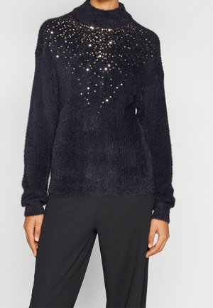 Kvinde iført en sort, blød sweater med spredte sølv-sekvenser på brystet kombineret med løse, sorte bukser.