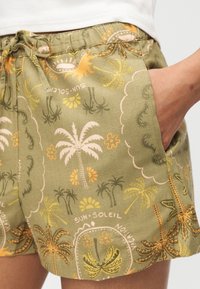 Personne portant un short vert orné d'un motif tropical de palmiers et de soleil, la main posée de façon décontractée dans la poche latérale, avec un haut blanc visible.