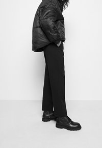 Osoba v černé péřové bundě, černých rovně střižených kalhotách a černých kožených šněrovacích botách na bílém pozadí.