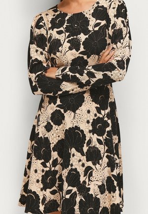 Beżowa sukienka z długimi rękawami, ozdobiona wyrazistymi czarnymi kwiatowymi wzorami, noszona przez osobę ze skrzyżowanymi rękami.