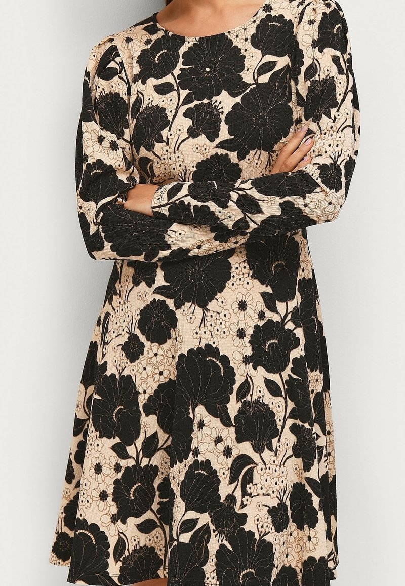 Robe beige à manches longues, ornée de motifs floraux noirs audacieux, portée par une personne les bras croisés.