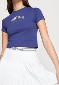Niebieski, bawełniany krótki T-shirt z logo "TOMMY JEANS" w białym i czerwonym kolorze. W połączeniu z białą spódnicą plisowaną, podkreśloną markowym paskiem w talii.