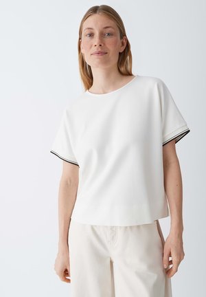 Femme aux cheveux blonds et raides portant un haut blanc à manches courtes avec un liseré noir et un pantalon clair sur un fond uni.