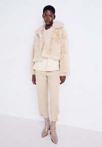 Veste en fausse fourrure beige avec une capuche, pull côtelé crémeux en dessous, pantalon beige et bottes brillantes beiges. Des boucles d'oreilles en argent ajoutent une touche de détail.