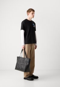 Sort t-shirt med logo, lagt over en hvid langærmet skjorte. Bred beige bukser og sort læder tote bag med præget "HUGO" logo.