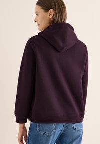 Sudadera de color púrpura oscuro con un bolsillo canguro y un corte relajado. Puños y dobladillo de canalé, textura suave, detalle de costura visible. Combinada con jeans azules.