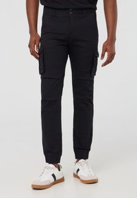 Czarne spodnie cargo mają wiele bocznych kieszeni, tapered fit, elastyczne mankiety i zapięcie na guzik w talii. Noszone z białymi trampkami.